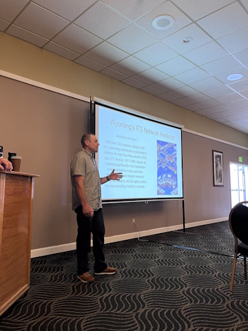A man gestures with his left hand while speaking beside a projector screen with slide text and image 'Wyoming's ITS Network Redesign.
